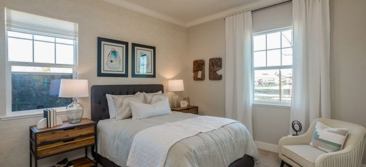 A cozy bedroom featuring a gray tufted headboard, soft bedding, and natural light coming through two large windows with white curtains, creating a calm ambiance.