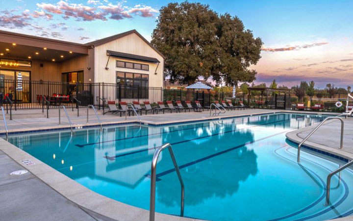 A clear blue swimming pool with lounge chairs arranged along the edge and umbrellas providing shade. The pool area is bordered by a fence, and the background features a modern building with warm interior lighting, set against a vibrant sunset sky.