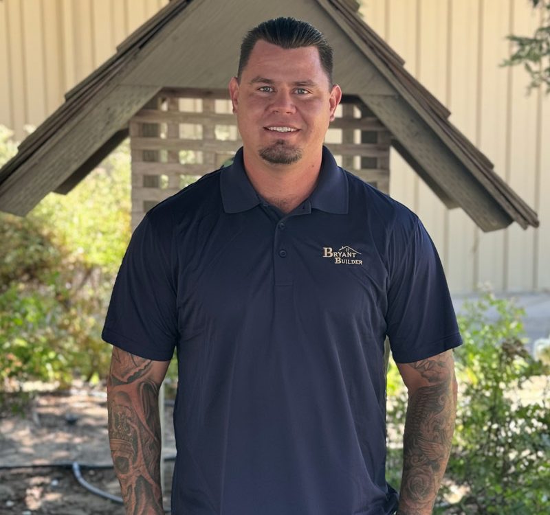 A professional portrait of the owner of Bryant Builders in a company polo shirt, standing in front of a gable roof structure.