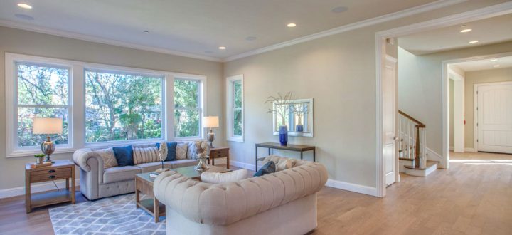 A large family room with light hardwood floors and large windows that provide a view of greenery outside, featuring tufted sofas and tasteful decor.