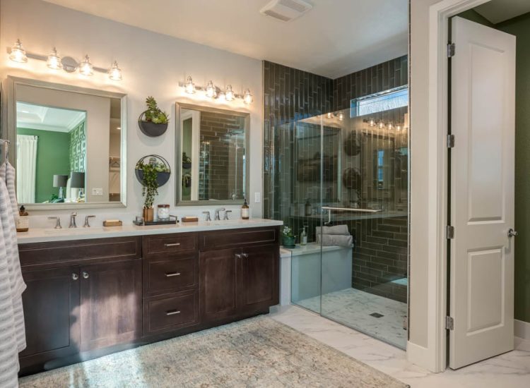 A spacious master bathroom featuring dual sinks, a dark tile walk-in shower, and elegant lighting fixtures above the mirror, with a view into the master bedroom.