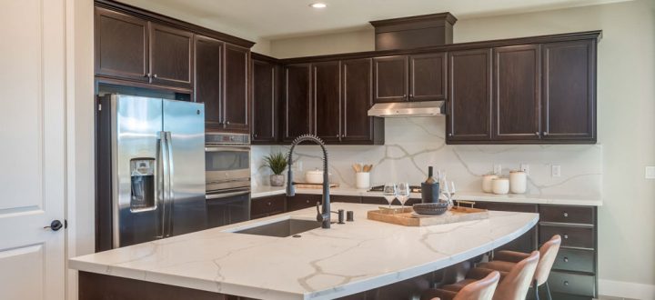 A sleek modern kitchen with dark cabinetry, marble countertops, and a central island with a large sink. The room includes stainless steel appliances and stylish leather bar stools.