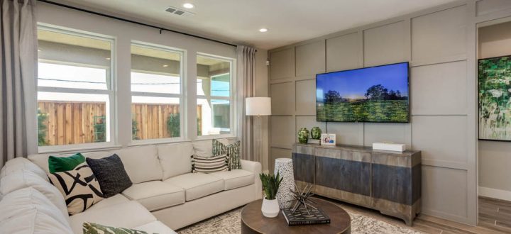A modern living room featuring a white sectional sofa, patterned pillows, and a large TV mounted on a sleek paneled wall, with large windows providing ample natural light.