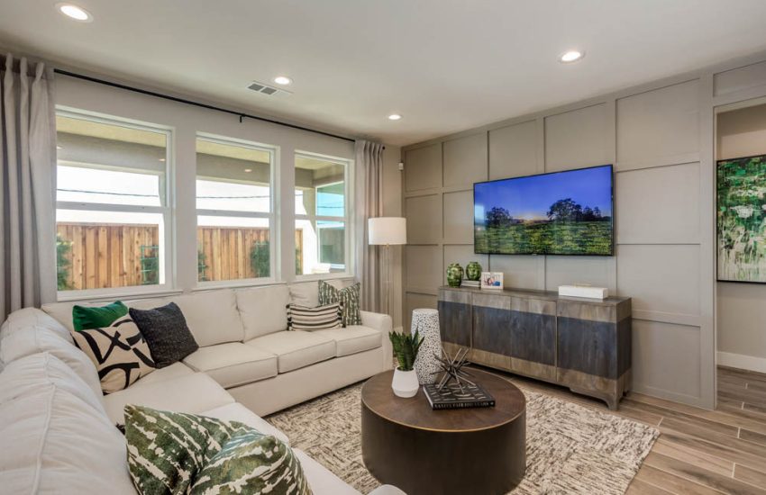 A modern living room featuring a white sectional sofa, patterned pillows, and a large TV mounted on a sleek paneled wall, with large windows providing ample natural light.