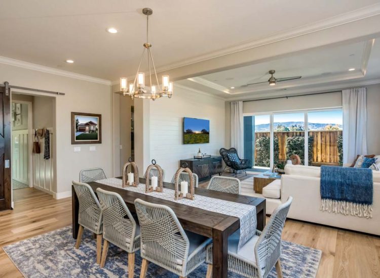 A cozy dining area with a rustic wooden table and chandelier, seamlessly flowing into an inviting living room with large windows that bring in plenty of natural light.