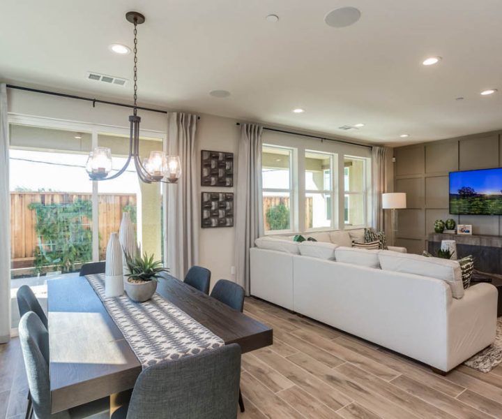 A bright open-plan dining and living area with a large wooden dining table, cushioned chairs, and a cozy sectional sofa. The space is flooded with natural light from large windows.