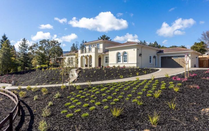 A single-story home with a beige exterior, multiple windows, and a landscaped front yard featuring mulched planting areas and shrubs. The property has a paved driveway and a clear blue sky in the background.