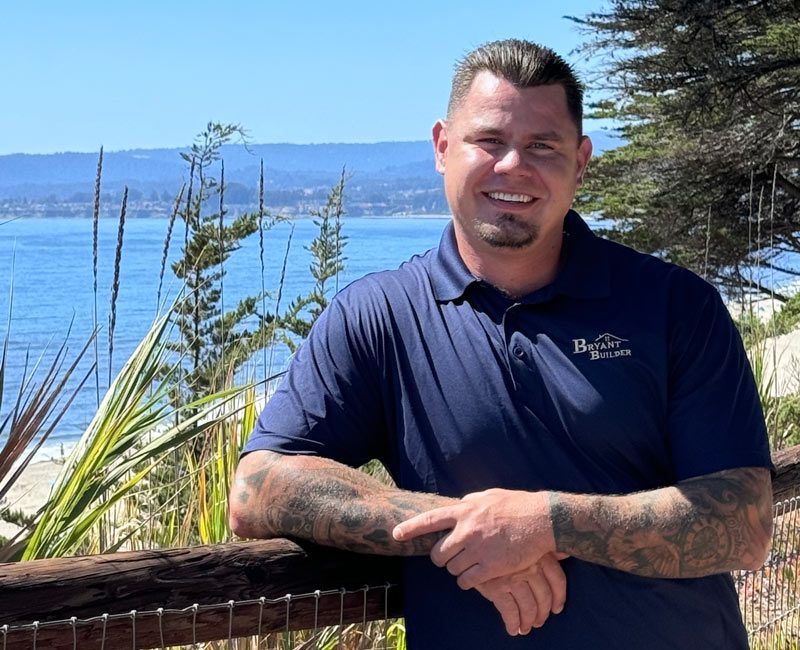 The owner of Bryant Builders, a middle-aged man with tattoos, smiling in an outdoor setting with a view of the sea in the background.